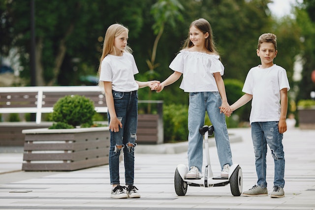 two children in blue jeans and white tee shirts help a third child ride a segway on a wooden path